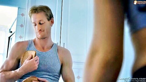 Muscular Caucasian man in tank top holds sandwich near woman's arm in blue top, teasing kitchen vibe
