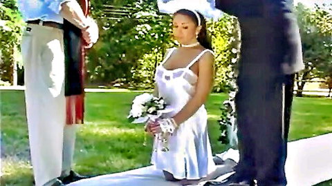 Brunette bride in revealing white slip dress kneels at outdoor wedding with two men