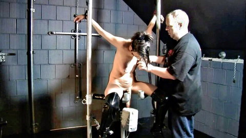 Clothed dominant man grips hair of bound naked submissive spread on BDSM metal frame