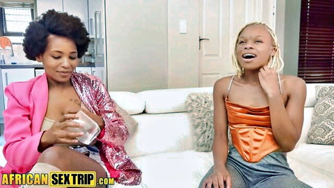 Two busty ebony women exposing tits on couch, one holding glass of white liquid provocatively