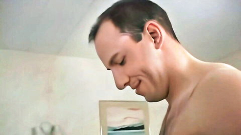 Shirtless man smiling while leaning over table examining small framed picture