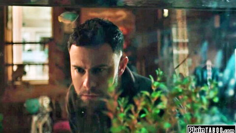 Bearded man in dark jacket peering intensely through green aquarium plants