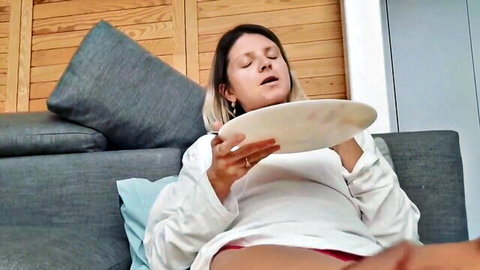 Brunette woman lounging on couch holds plate of cum with eyes closed in pleasure