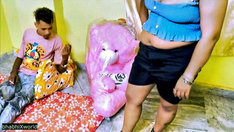 Chubby Indian wife with big tits and fat belly in blue frilly top and black shorts stands by sitting husband and pink teddy bear