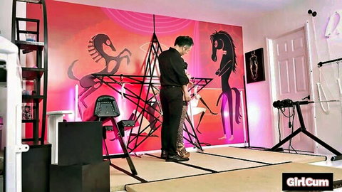 Brunette woman in tight black outfit stands in BDSM dungeon with bondage chair and toys