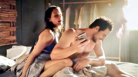 Brunette in tank top spoons with shirtless muscular hunk on bed holding phone smiling playfully