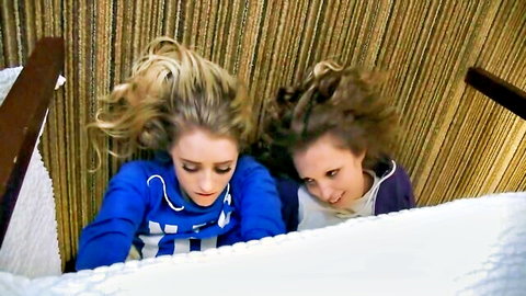 Blonde and brunette girls peeking playfully from bed edge in hoodies