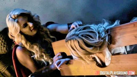 Blonde dominatrix in red costume holds black ball gag to submissive blonde's mouth bent over wooden bench