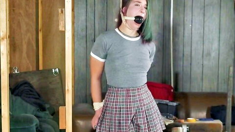 Ball-gagged schoolgirl slave with wrists bound standing in bondage garage scene