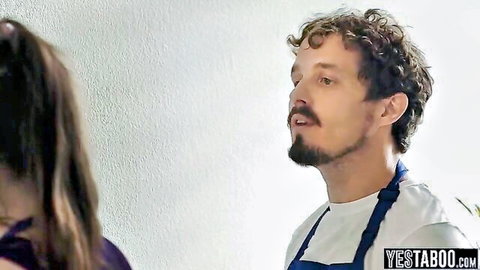 Bearded curly-haired chef in apron gazes at seductive brunette woman in kitchen tease