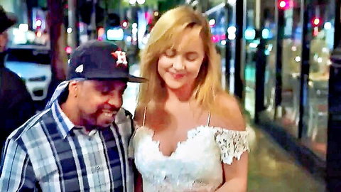 Blonde in low-cut white lace dress smiles with mature black man in Astros cap on neon-lit street