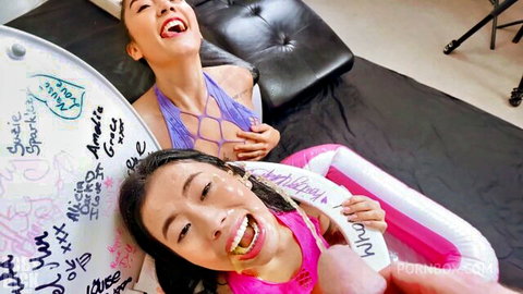 Two Asian sluts grinning with yellow cum on teeth as thick cock hovers near mouth beside signed pink floatie