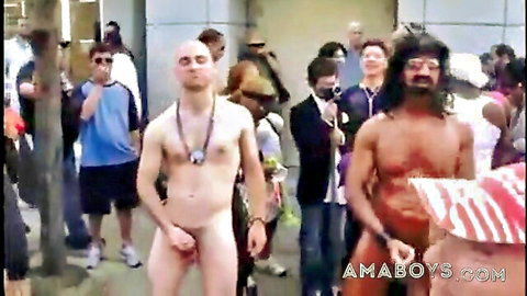 Two muscular naked men stand nude with hands covering cocks amid public outdoor crowd