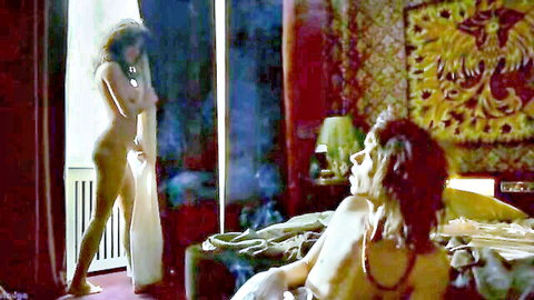 Nude curvy woman covers her tits standing by window as naked man lounges on bed staring