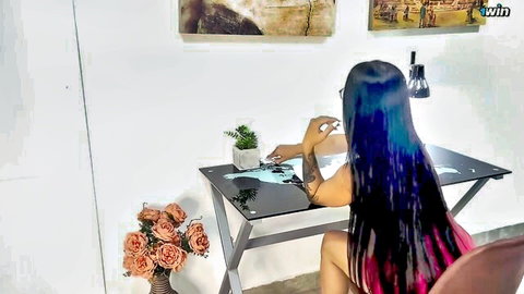 Blue-haired tattooed Asian woman in black top at desk, teasing pose