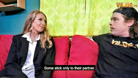 Blonde MILF in white blouse and black suit chats closely with long-haired young guy on red couch during casting interview