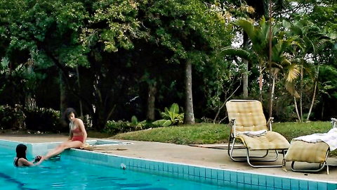 Two bikini-clad babes lounge poolside, brunette in red swimsuit dips legs in water while friend swims in black bikini