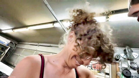 Curly haired MILF in glasses smiles with head thrown back wearing red tank top in garage workshop