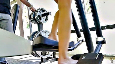 Athletic fair-skinned woman spreads bare legs wide on leg press machine in gym with black trainer
