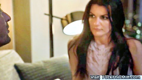 Brunette MILF in low-cut dress sits closely with man on couch teasing with cleavage