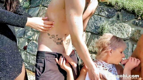 Blonde short-haired babe kneels outdoors to suck tattooed hunk's cock while women grope his body