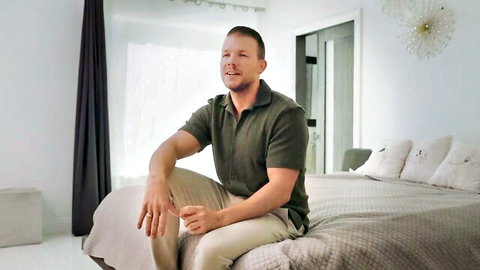 Muscular white man in green polo shirt sitting smiling on bedroom bed