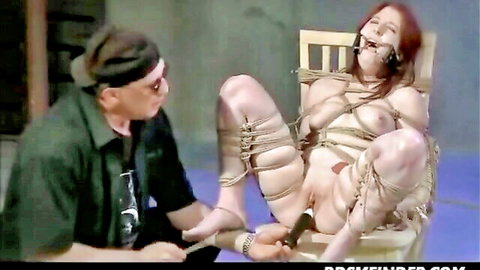Gagged redhead submissive bound naked to chair with legs spread exposing shaved pussy as dom holds cane to her cunt