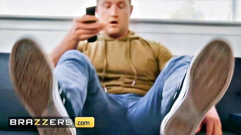 Shocked young guy in hoodie and jeans spreads legs wide on couch holding phone in Brazzers scene
