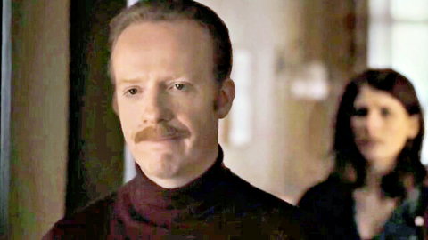Red-haired man with mustache in turtleneck stares intently with brunette woman blurred in background