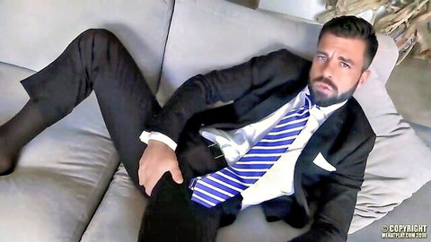Hector de Silva in suit, hand in pants, lounging seductively on couch