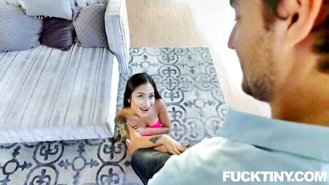 Petite Asian teen on knees smiling up at man's crotch with deep cleavage in pink top