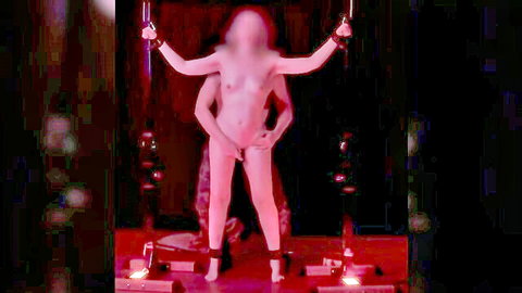 Petite nude fair-skinned woman with small tits arms raised cuffed to poles legs spread exposing pussy in red-lit bondage stage