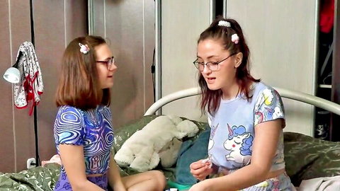 Two cute brunette teens with glasses chatting intimately on bed in casual tops