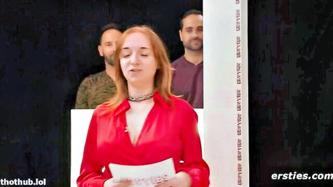 Redhead in deep cleavage red blouse speaks on stage with bearded and dark-haired men behind her