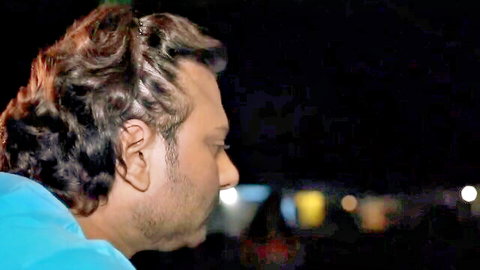 Profile of curly-haired South Asian man with beard in blue shirt looking sideways at night