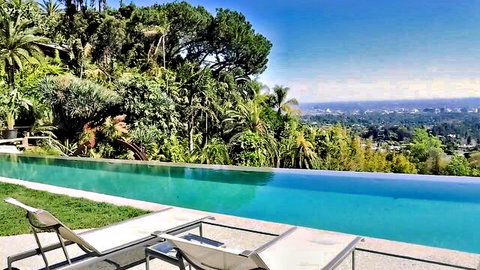 Turquoise infinity pool with white lounge chairs overlooking lush Hollywood Hills and city skyline