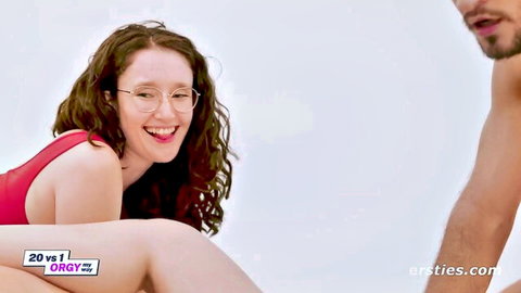 Smiling curly-haired brunette with glasses in red top kneels with shirtless man in amateur gangbang tease