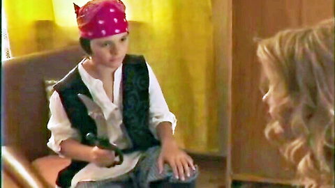 Young boy in pink bandana pirate costume with toy sword sits on couch chatting with blonde girl
