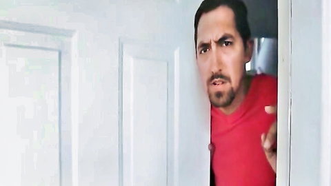 Suspicious Latino man with beard peeking intensely from behind white door in red shirt