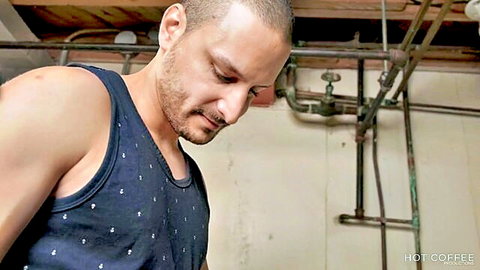 Muscular shaved-head man in tight tank top looking down intently in industrial basement