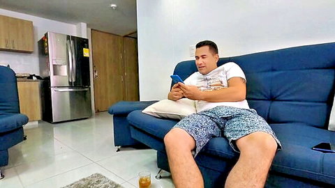 Handsome tanned man in shorts and t-shirt sitting on blue couch holding phone in modern living room
