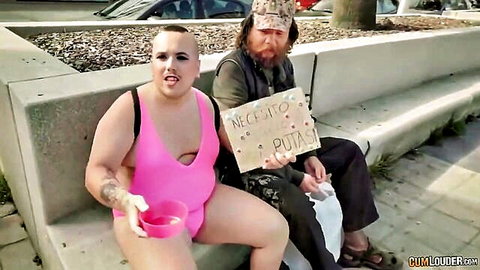 Drag queen in skimpy pink bikini holds cum bowl beside homeless man with 'Necesito putas' sign