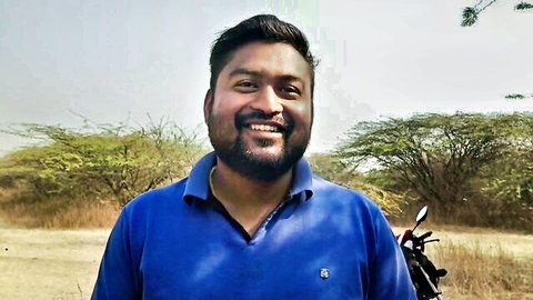 Smiling bearded Indian man in blue polo shirt posing outdoors with motorcycle in jungle background