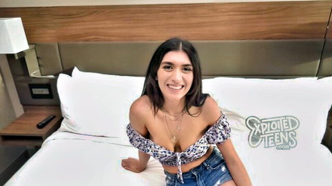 Smiling busty teen with huge tits in low-cut off-shoulder top teasing on hotel bed