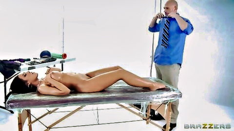 Nude oiled brunette with big fake tits lies spread on massage table as photographer captures her