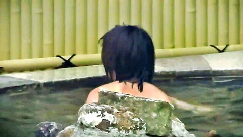 Asian woman with black hair relaxing topless in outdoor hot spring bath from behind