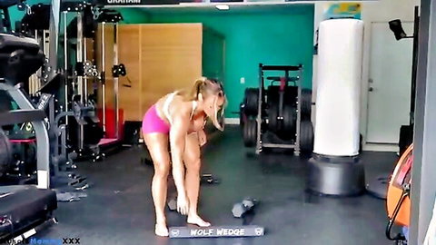 Fit woman in pink shorts bending over for hamstring dumbbell workout in gym