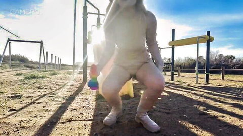 Babe in tight bodysuit squats in sunny public park playground holding rainbow dildo at her crotch for toy insertion