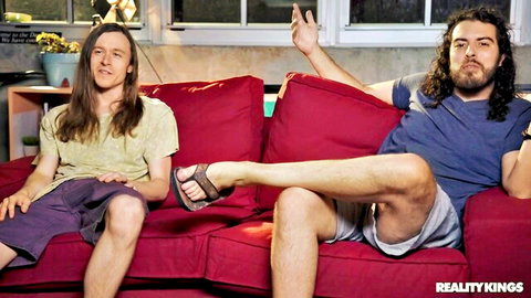 Two long-haired guys lounging casually on red couch in living room with flip-flops and shorts