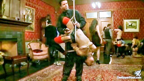 BDSM party with suspended male slave in red ropes held by suited master